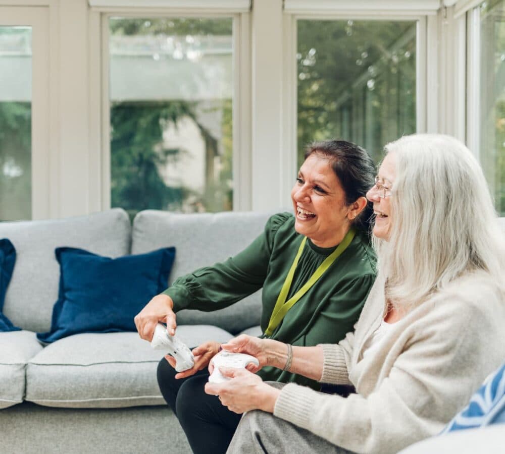 An older female adult with long grey hair sitting on a couch with her younger female carer with long black hair and wearing green both happy playing together inside the house