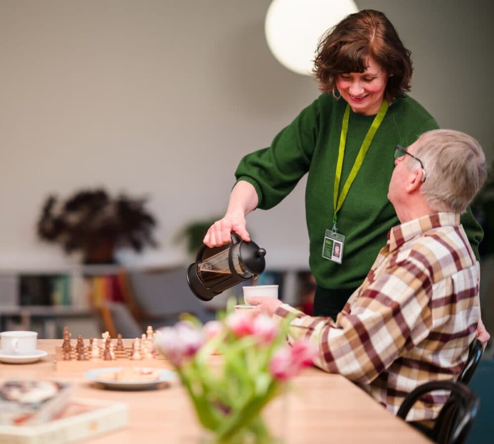 A female carer wearing green pouring coffee to an older male adult with grey hair and wearing eyeglasses while sitting on the table