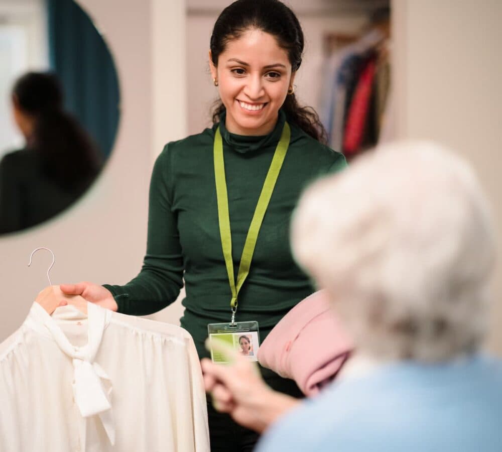 An older female adult with white hair being helped by her younger carer wearing green and with long hair holding her clothes to wear inside the room