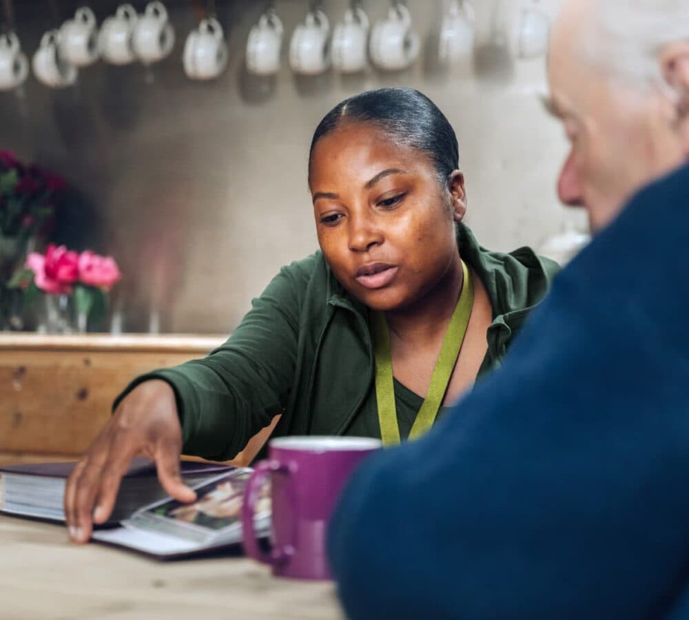 A woman shows a book to an older man at a table, with mugs and cups in the background. - Home Instead