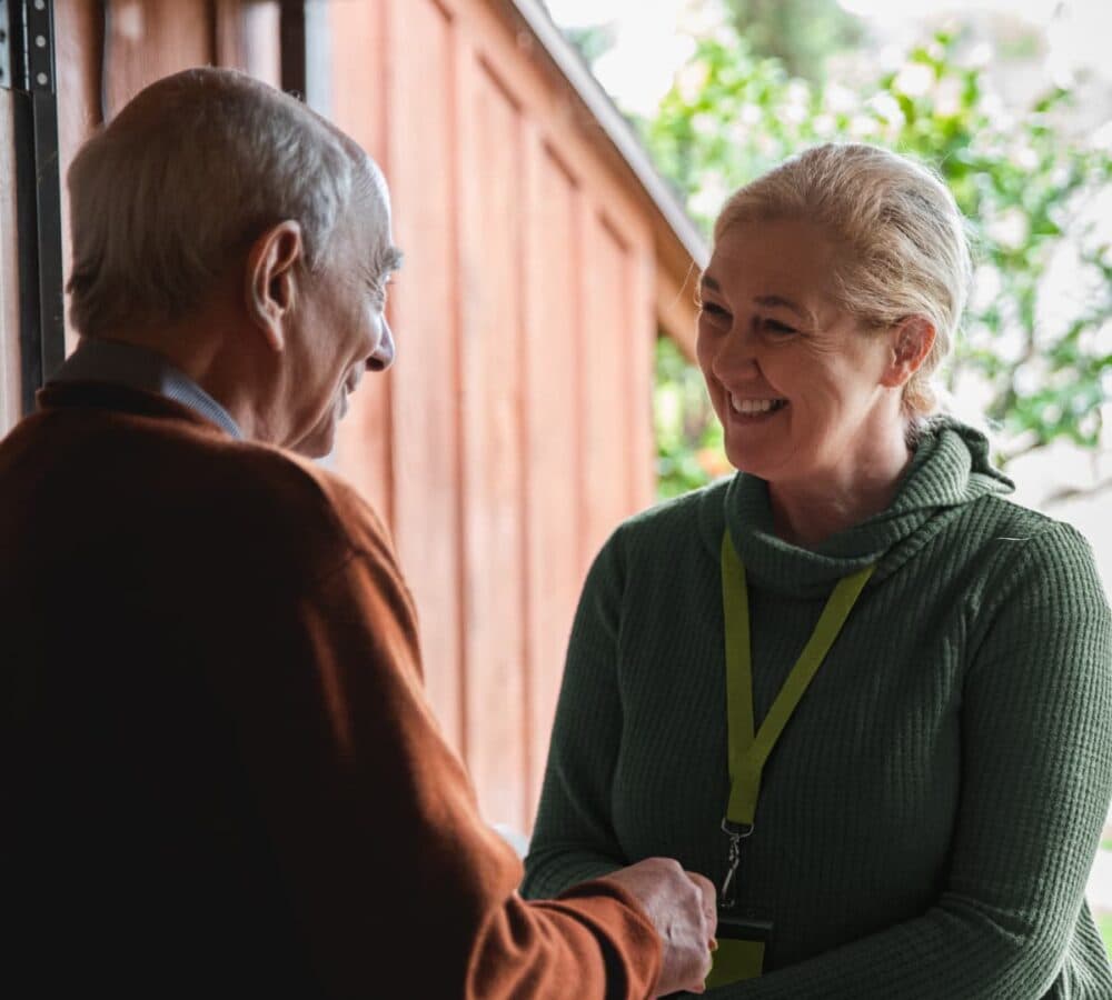 An older male adult with grey hair at the door welcoming his female carer wearing green and happy and smiling while shaking hands
