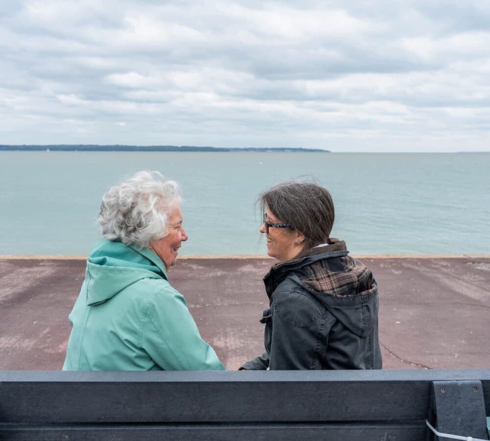 An older female adult with white hair wearing green coat sitting by the beach with her younger female carer wearing eyeglasses and with long hair