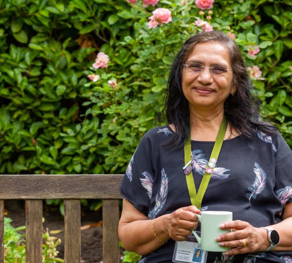 A woman with long black hair and wearing eyeglasse and black top happy and smiling while holding a cup of coffee and sitting on a bench with flowers and plants at her back