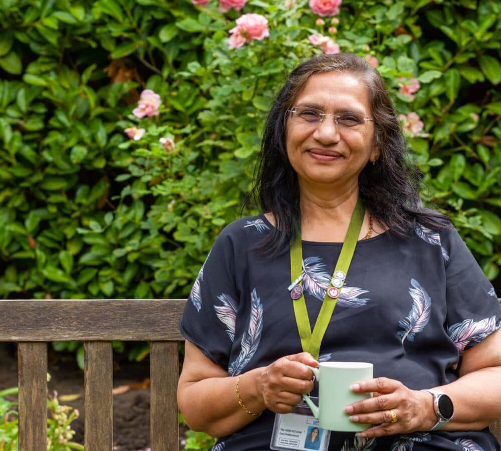 Smiling woman with glasses sits on a bench holding a mug, with flowers and greenery in the background. - Home Instead