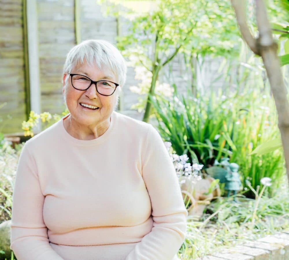 An older female adult with white hair wearing eyeglasses smiling and wearing long sleeves in the garden