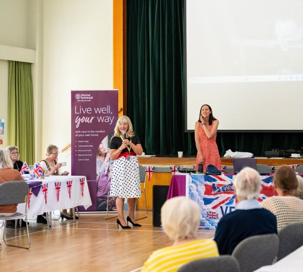 Two women speak on stage at a community event; people sit and watch in a decorated hall. - Home Instead