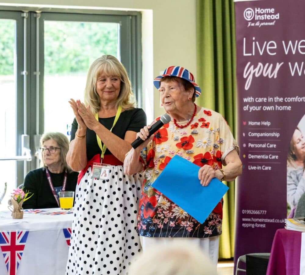 Two women stand together, one holding a microphone and papers, at a community event with care service banners. - Home Instead