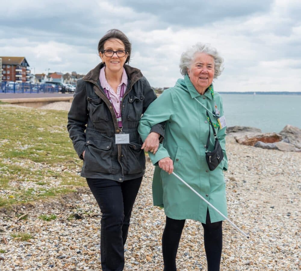An older female adult with white hair wearing green coat walking by the beach and using a crane with her younger female carer wearing eyeglasses and with long hair