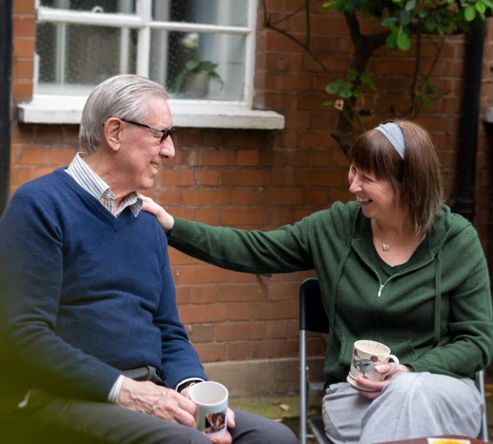 An older man and a woman sit outside, smiling and holding mugs, as the woman rests her hand on his shoulder. - Home Instead