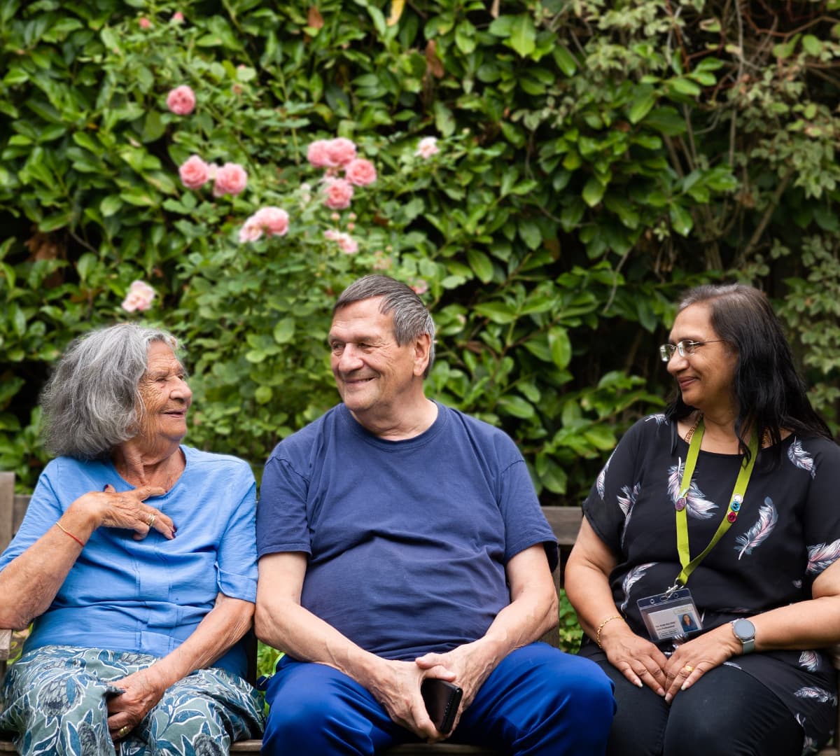 An older female adult with grey hair and wearing blue top smiling with her husband with grey hair and wearing blue and with their younger female carer with black long hair and wearing eyeglasses while they are all sitting on a bench happy and smiling in the garden