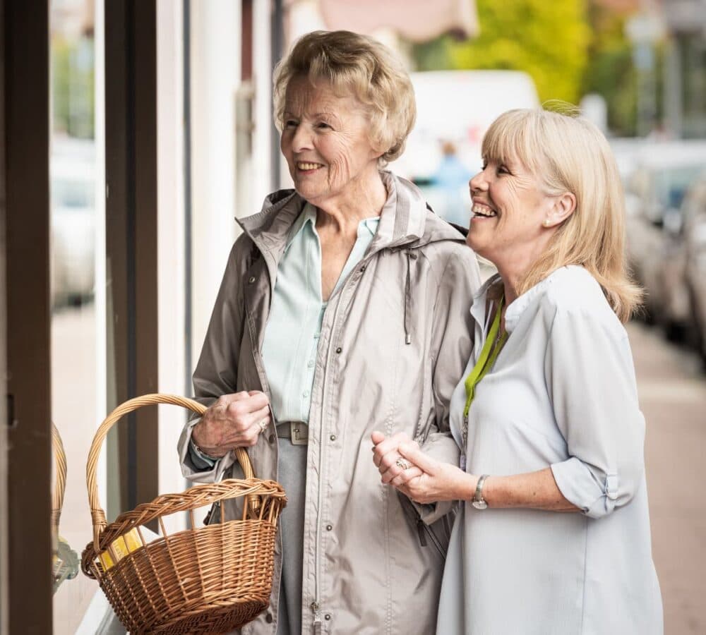 An older female adult with short hair and wearing a jacket and bringing a basket with her younger female carer with blonde hair both smiling outdoors