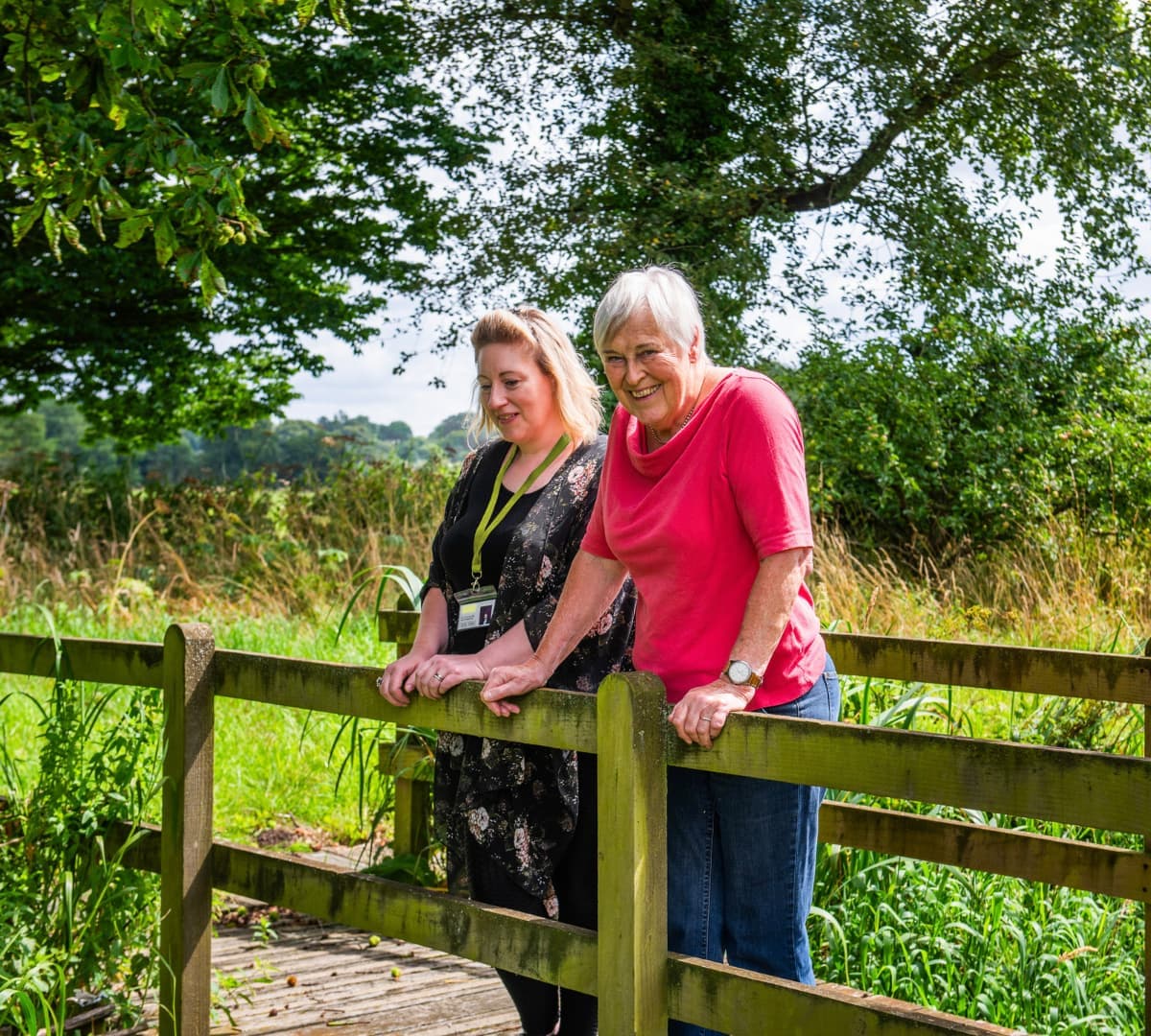 An older female adult on the bridge and wearing pink wth short hair accompanied by her younger female carer with black dress both happy and smiling