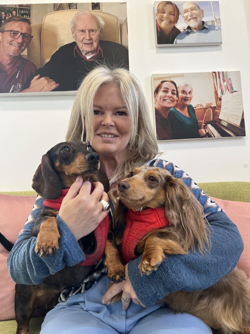 Smiling woman holding two dachshunds in red harnesses, seated on a couch with photos on the wall behind her. - Home Instead