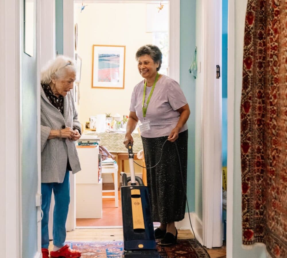 An older female adult with white hair chatting with her younger female carer wearing skit and vacuuming the floor of the house while smiling