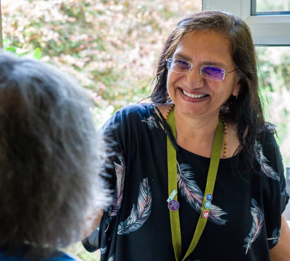 A female carer wearing eyeglasses and with long black hair smiling at the door of the house while beoing welcomed by an older female adult