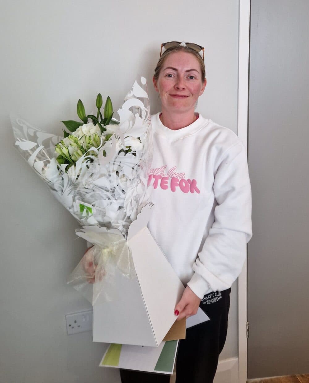 Woman in a white sweatshirt holding a bouquet of white flowers and some cards, standing indoors. - Home Instead