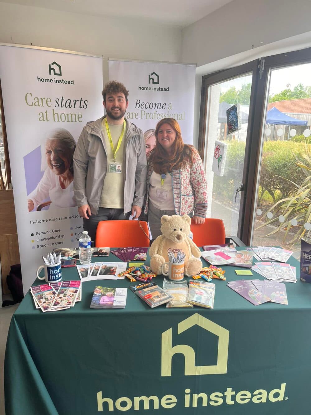 Two people stand behind a "home instead" care stall with brochures, a teddy bear, and info materials on a table. - Home Instead