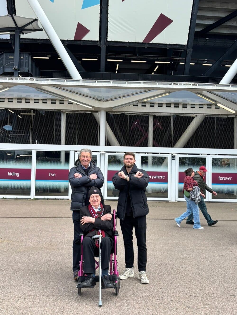 Three men pose with arms crossed in front of a stadium, one is seated in a wheelchair. - Home Instead