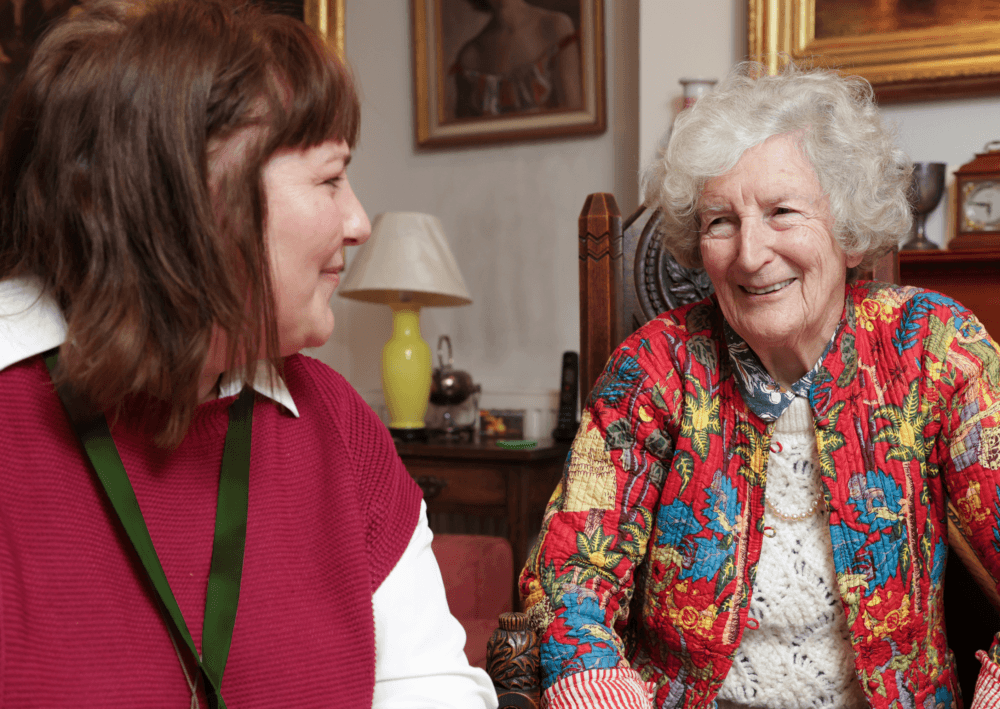 Two women smiling and talking together in a cozy, warmly decorated room. - Home Instead