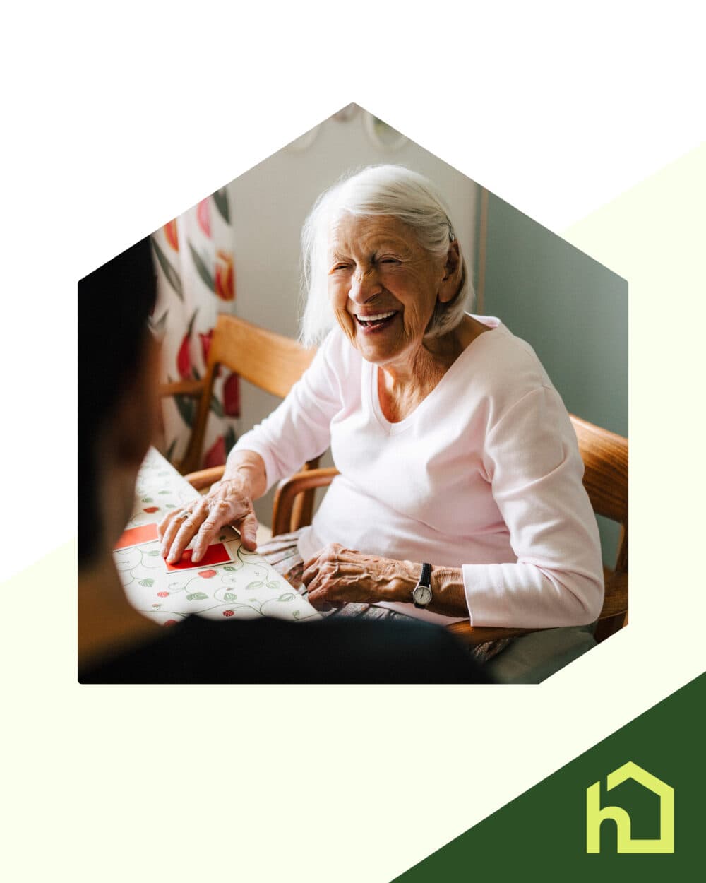 Smiling elderly woman sitting at a table, talking to someone, with a green house logo in the corner. - Home Instead