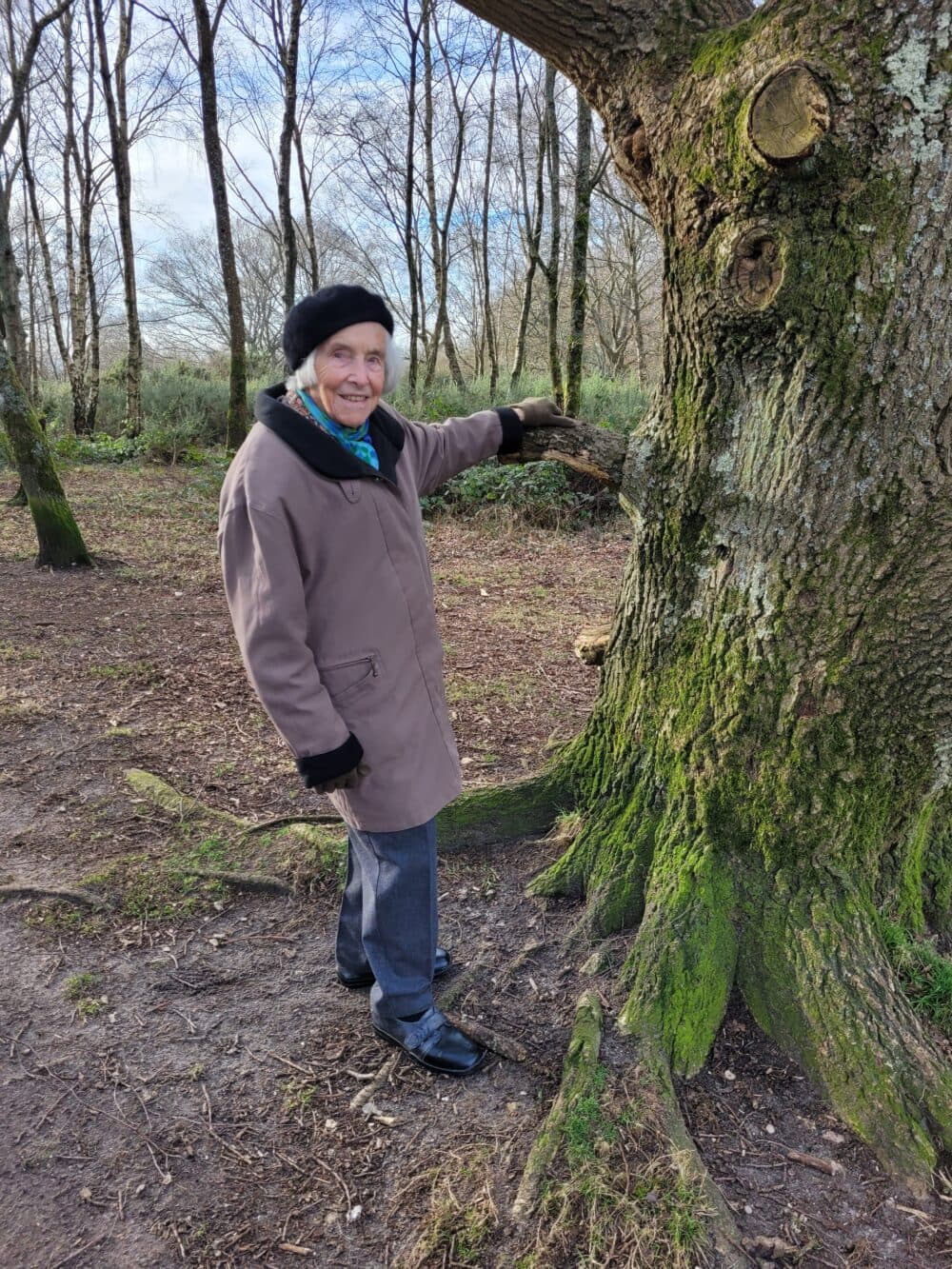 Smiling elderly person in a coat and beret touching a large tree in a wooded park. - Home Instead