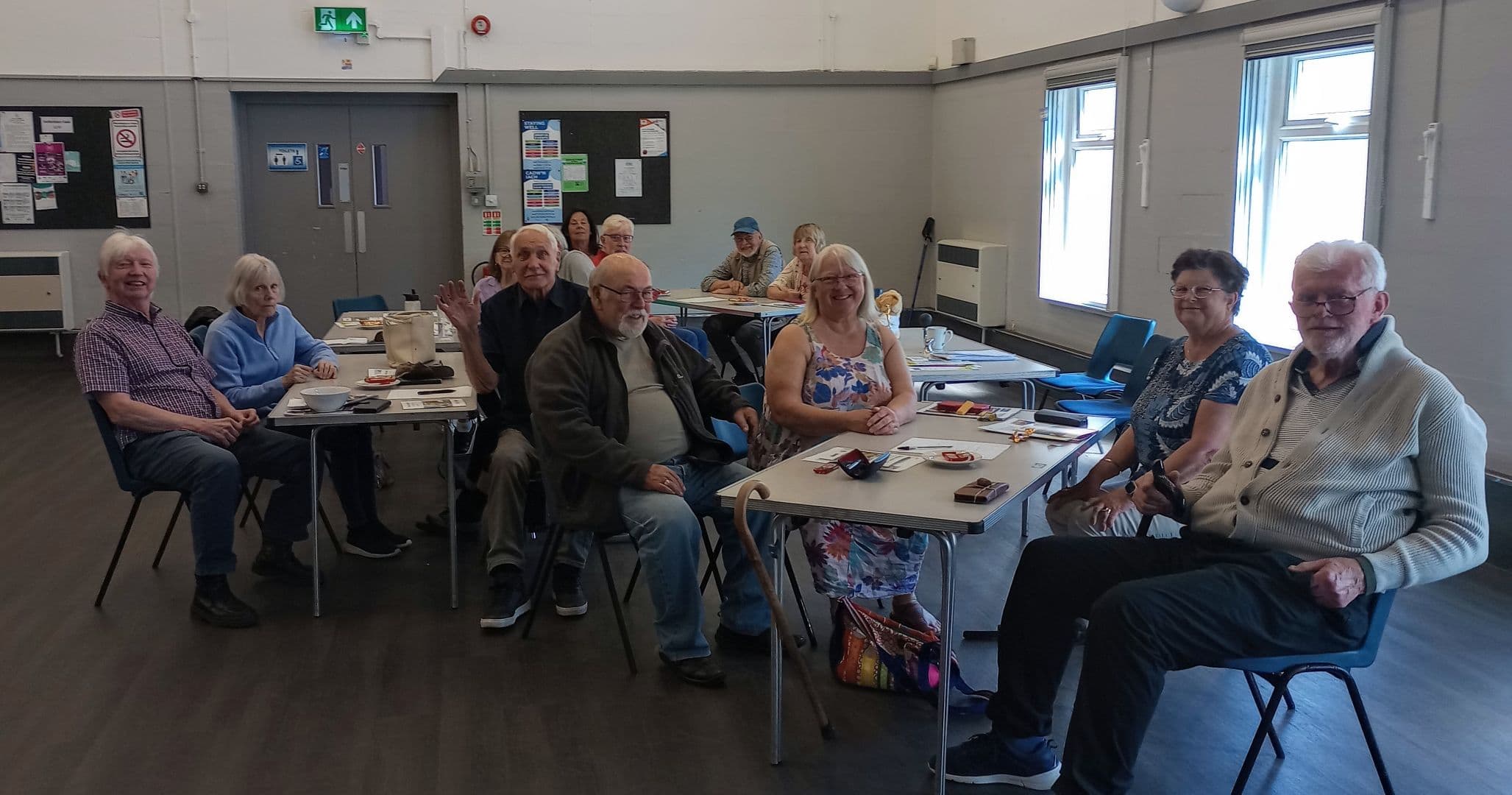 A group of older adults sit at tables in a bright room, smiling at the camera. - Home Instead