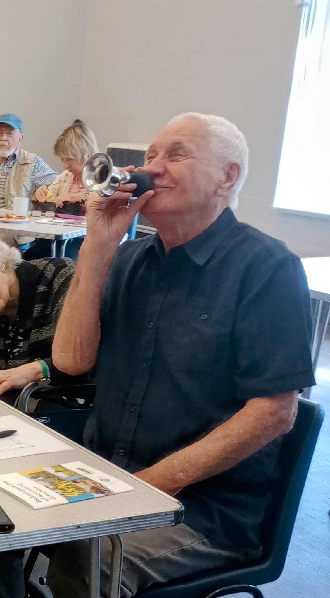 Elderly man smiling and holding a small horn to his mouth while sitting at a table in a bright room. - Home Instead