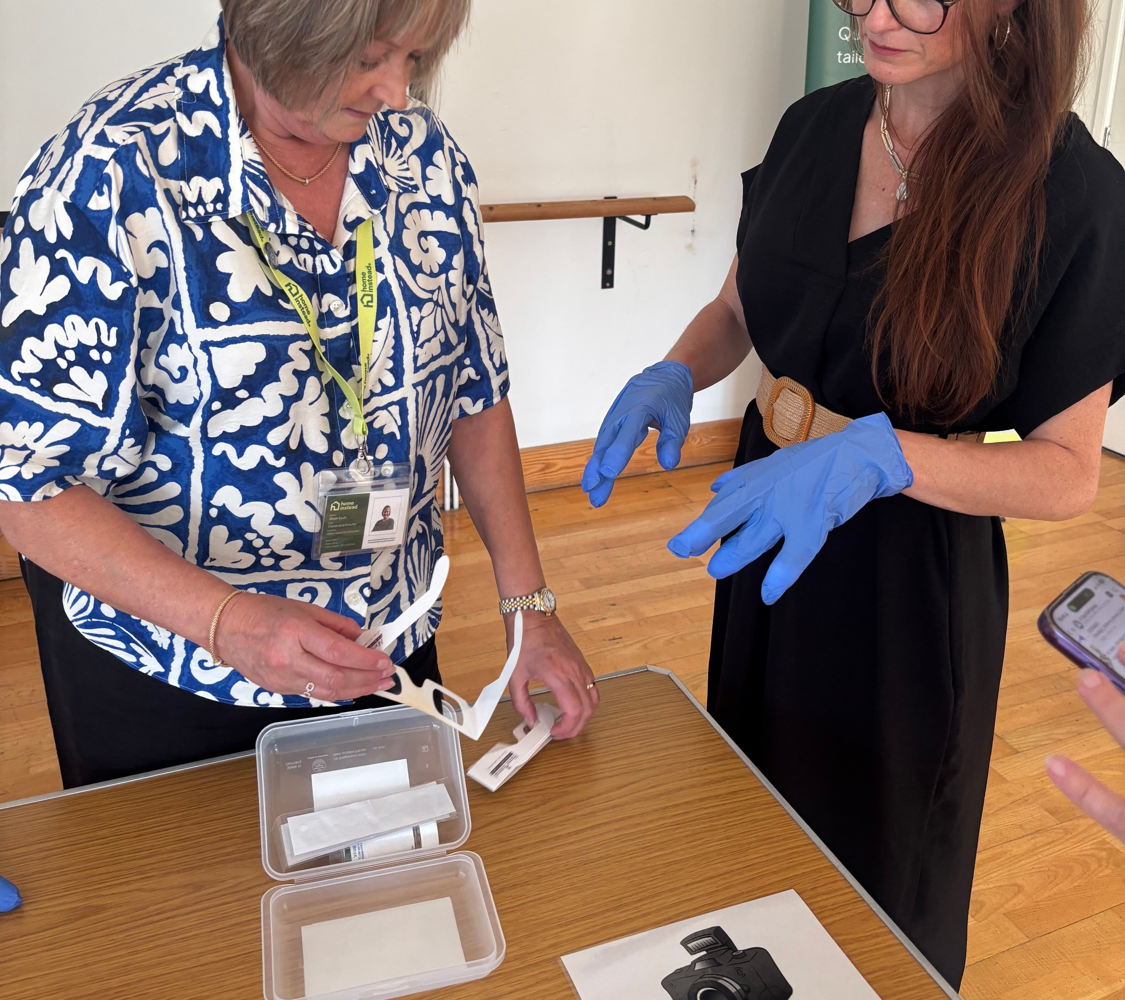 Two women at a wooden table during a healthcare event. One woman in a blue and white patterned shirt with a lanyard is handling medical testing equipment from clear plastic containers, while another woman in a black top wearing blue medical gloves observes. A green banner with 'Care' and a house icon is visible in the background, along with a sign mentioning photography consent.