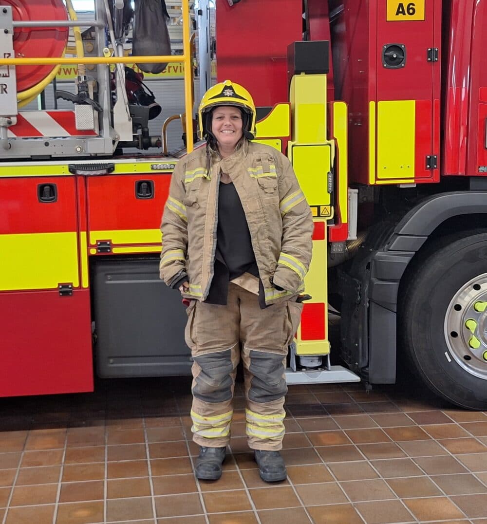 A smiling firefighter in full gear stands in front of a red fire truck indoors. - Home Instead