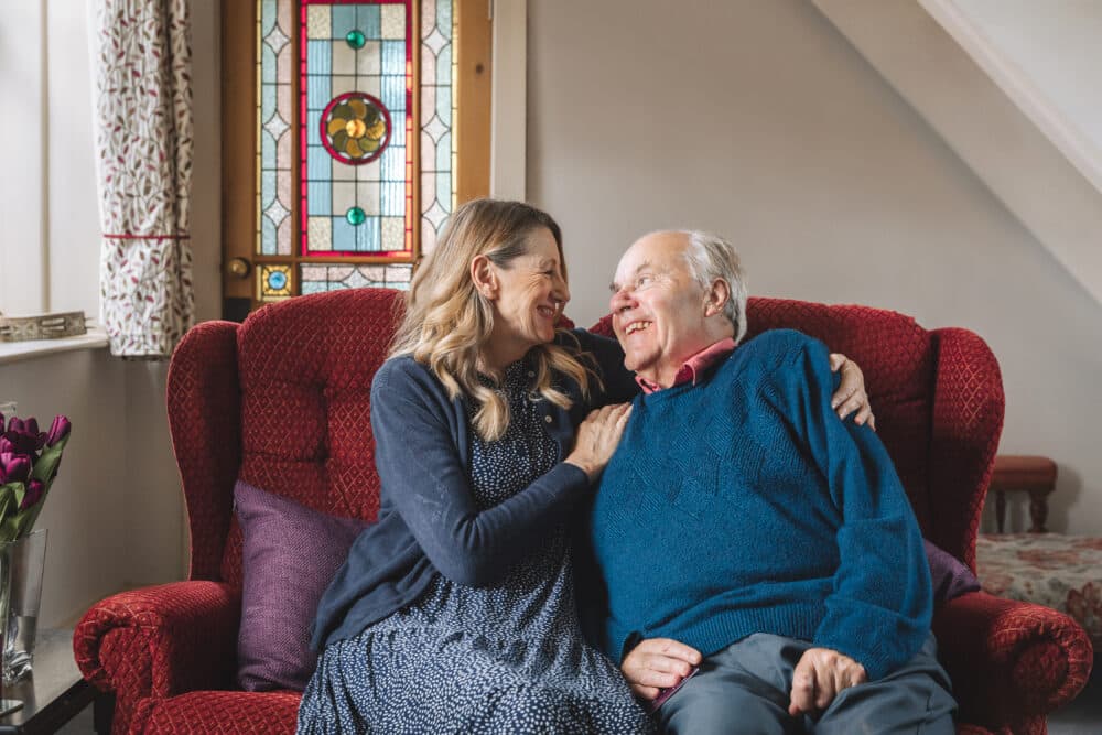 A smiling woman and elderly man sit close together on a red sofa, sharing a joyful moment at home. - Home Instead