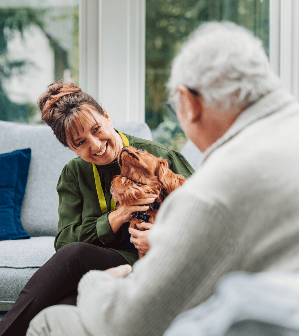 Smiling woman with a dog sits on a sofa, facing an elderly man in a light sweater. - Home Instead
