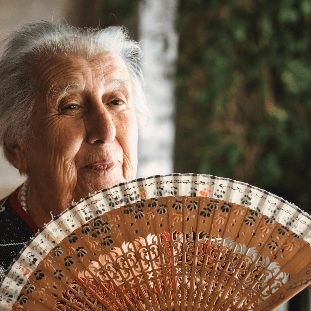 Elderly woman smiling gently, holding an ornate fan, with a leafy background. - Home Instead