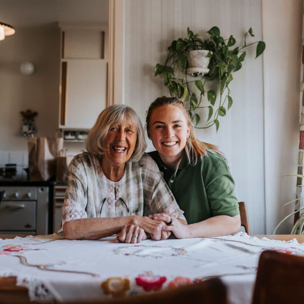 An elderly woman and a young woman smile, sitting together at a table with a floral tablecloth. - Home Instead