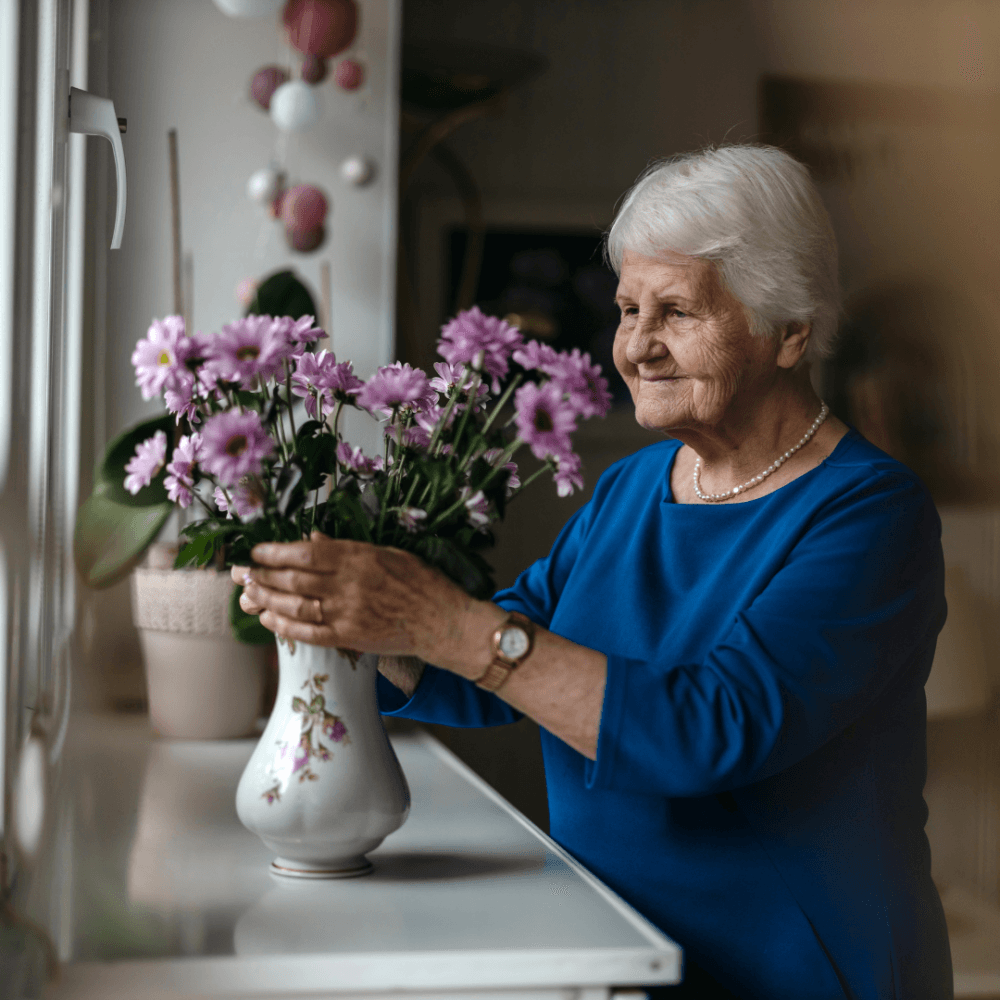 Elderly woman in a blue dress arranging pink flowers in a vase by a window. - Home Instead