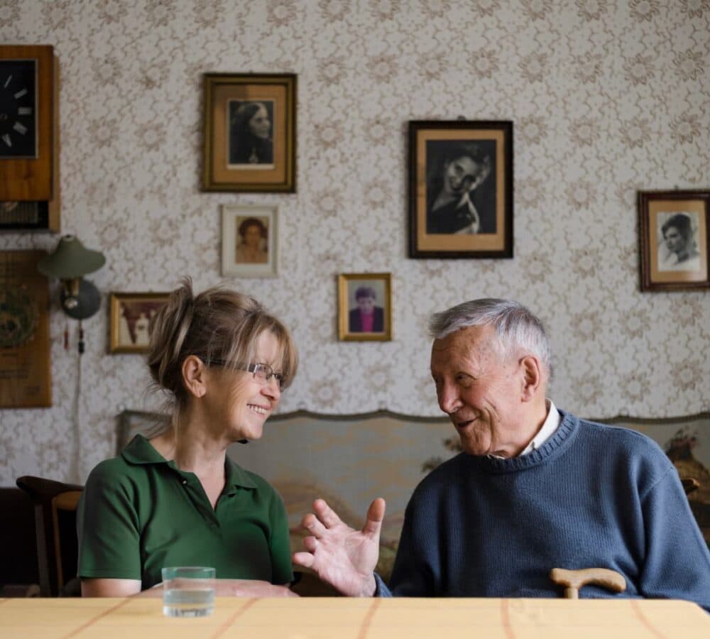 An older male adult with grey hair wearing blue pullover chatting with his female carer with brown hair and wearing green both smiling and sitting on a chair inside the house