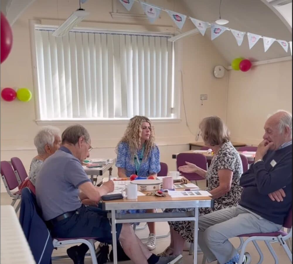 An older female adult with white hair and wearing green sweater with her younger female carer holding a puppy while chatting both happy and smiling