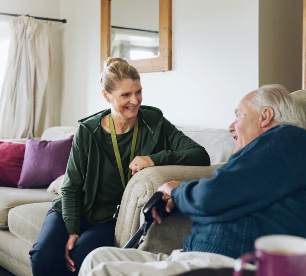 A caregiver smiles while talking to an elderly man sitting on a sofa in a cozy living room. - Home Instead