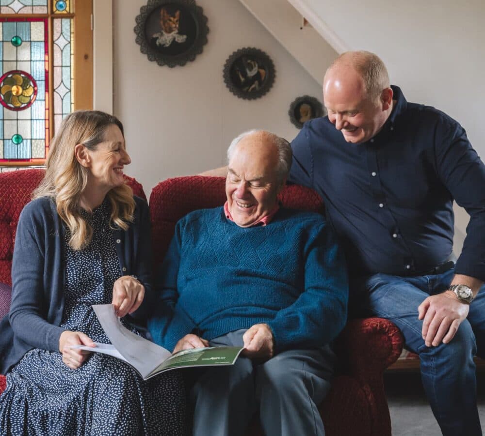 Three people sit on a red couch, smiling and looking at a booklet together in a cozy living room. - Home Instead