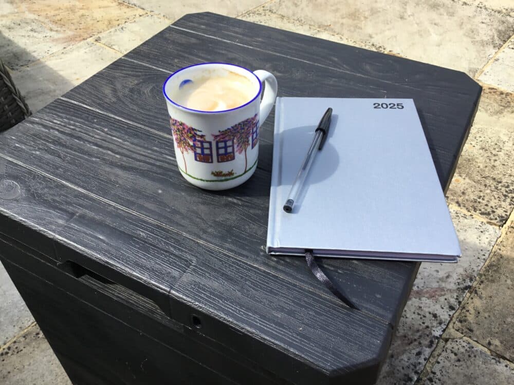 A coffee mug, a 2025 planner, and a pen on a black outdoor table with stone paving. - Home Instead