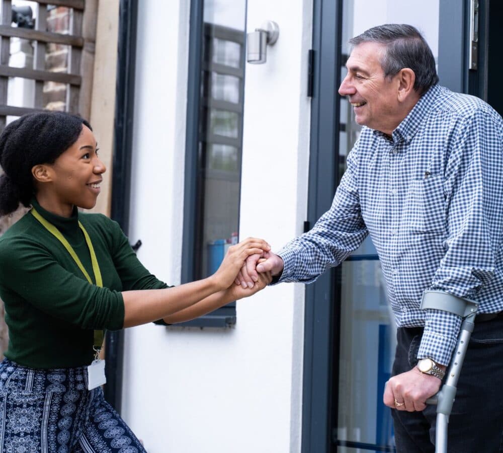 An older male adult with grey hair and wearing log sleeves and using a crutch going out of the house with his younger female carer with black curly hair, wearing green smiling and helping him while going out of the house