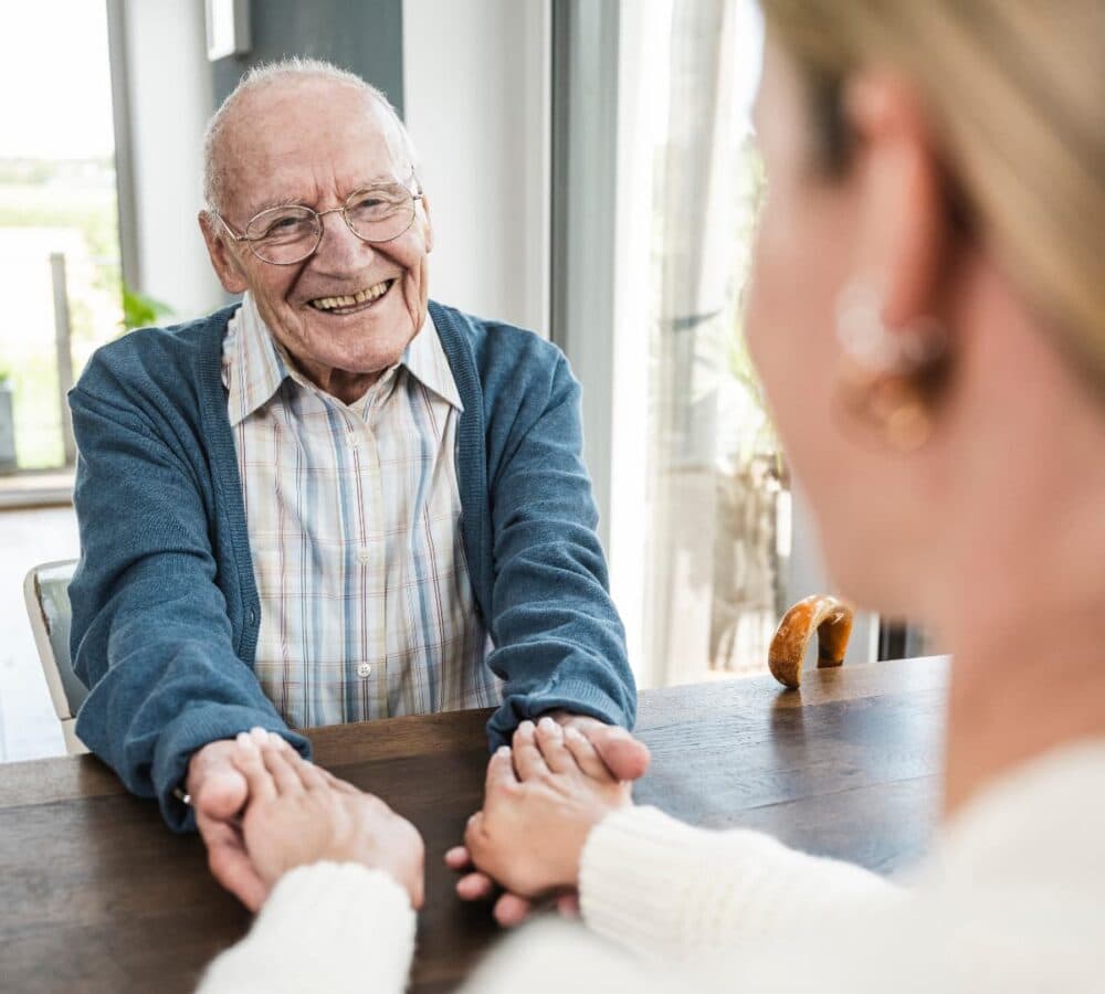 Smiling elderly man holding hands with a woman across a table in a bright, cozy room. - Home Instead