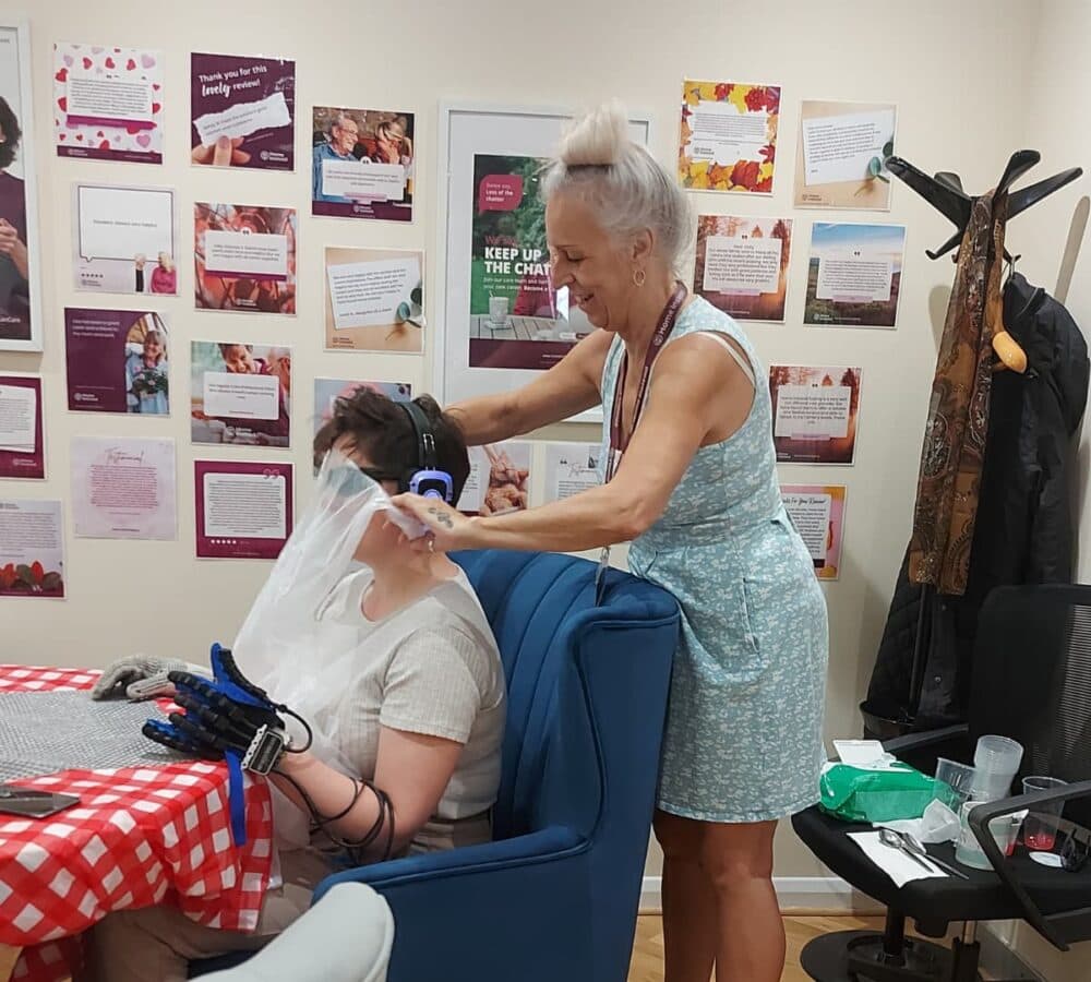 A woman helps another person wearing gloves and headphones with a cloth in a room decorated with posters. - Home Instead