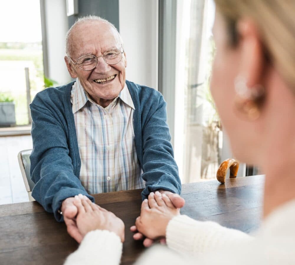 An older male adult wearing eyeglasses and sweater smiling while holding the hand of his daughter inside the house