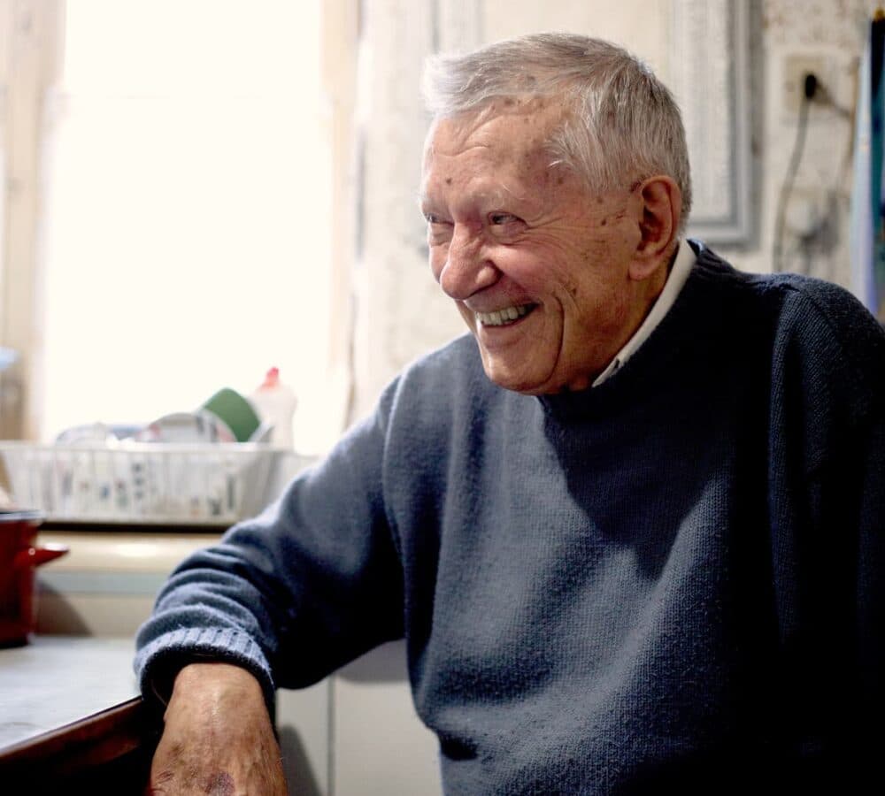Smiling elderly man in a gray sweater sits at a kitchen table with sunlight coming through the window. - Home Instead