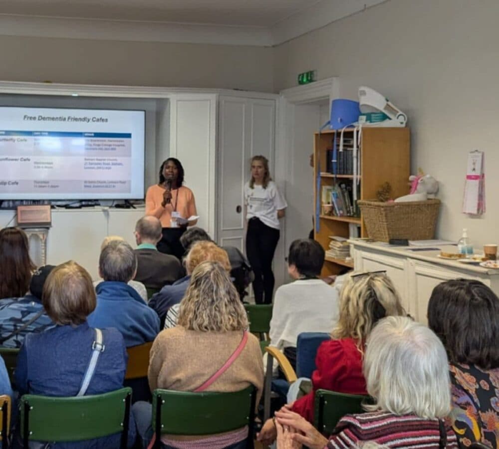 A woman speaks at a dementia-friendly event to an audience, with a slideshow displayed behind her. - Home Instead