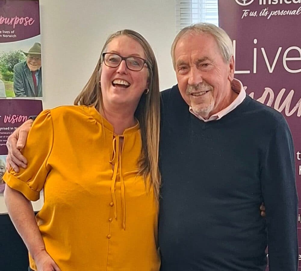 Two people smiling and posing together indoors in front of purple posters. - Home Instead