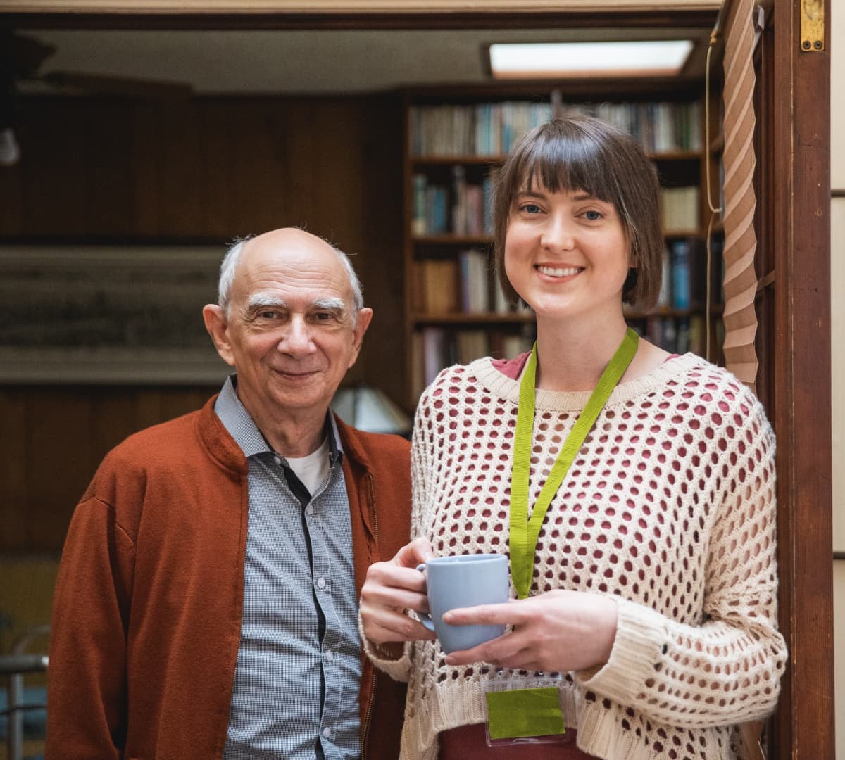 An older male adult with white hair and wearing a sweater with his younger female carer with short hair and holding a cup of coffee both smiling and standing inside the house