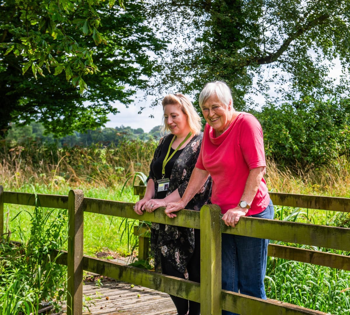 An older female adult with grey hair and wearing fuschia pink standing on the bridge with her younger female carer with short hair and wearing black both happy and smiling
