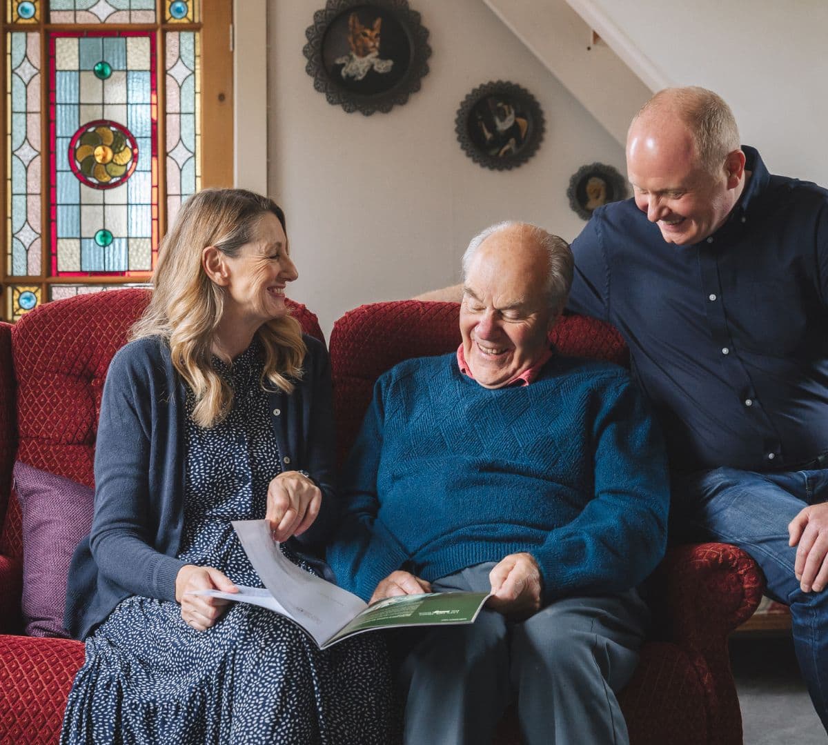 Three people sit on a red couch, smiling and looking at a book together in a cozy living room. - Home Instead