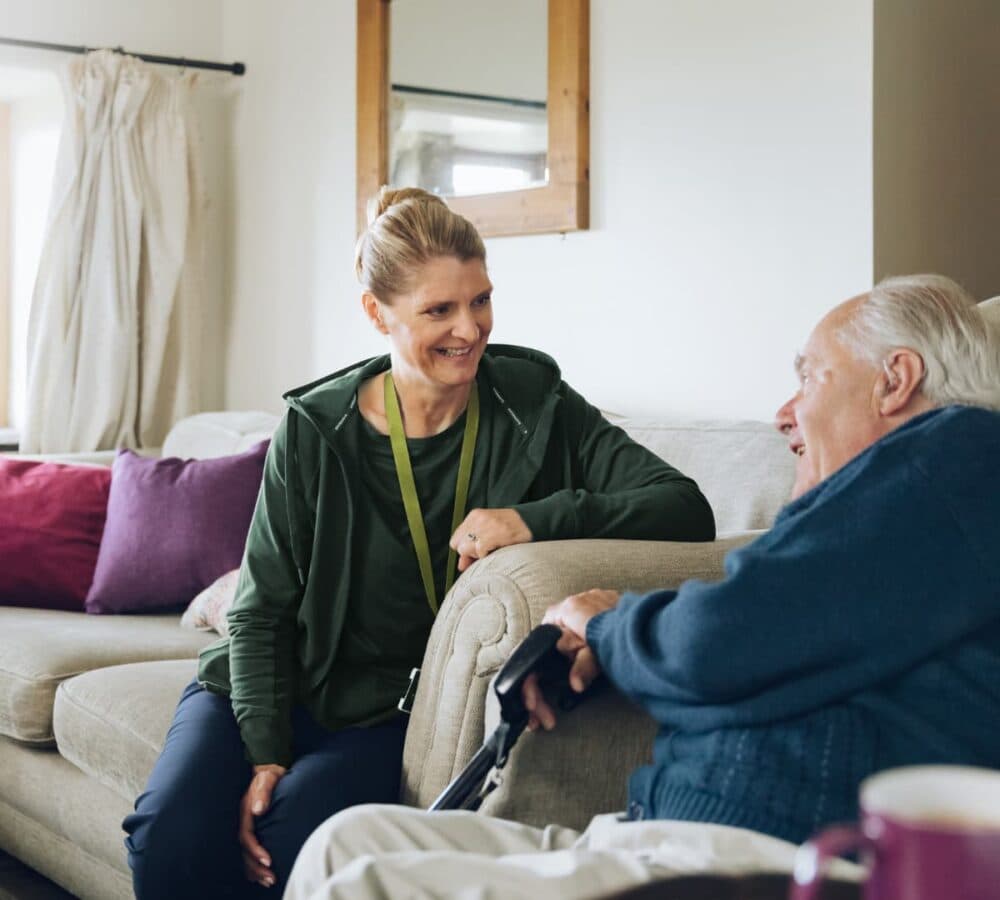 A woman sits and smiles, talking with an elderly man holding a cane on a sofa in a cozy living room. - Home Instead